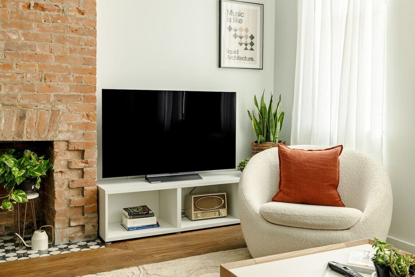 A living room with a brick fireplace, a flat-screen TV on a white stand, a white armchair with a brown cushion, and a framed quote on the wall.