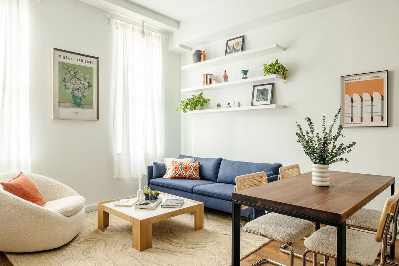 Ein modernes Wohnzimmer mit einem blauen Sofa, einem weißen Sessel, einem Holztisch und einem Esstisch mit Stühlen. Regale mit Pflanzen und Büchern sowie gerahmte Kunstwerke zieren die Wände.