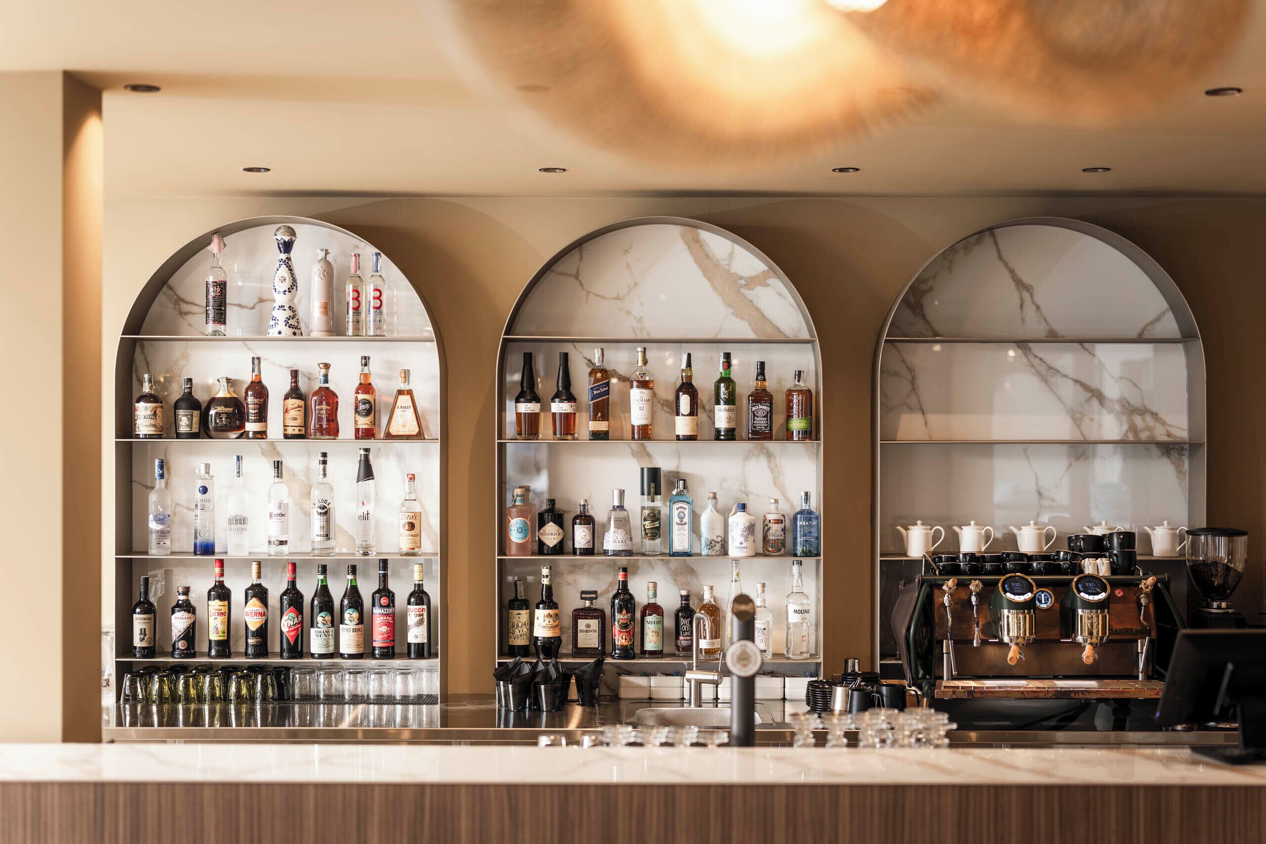 A bar area with shelves of liquor bottles, a coffee machine, and a marble wall.