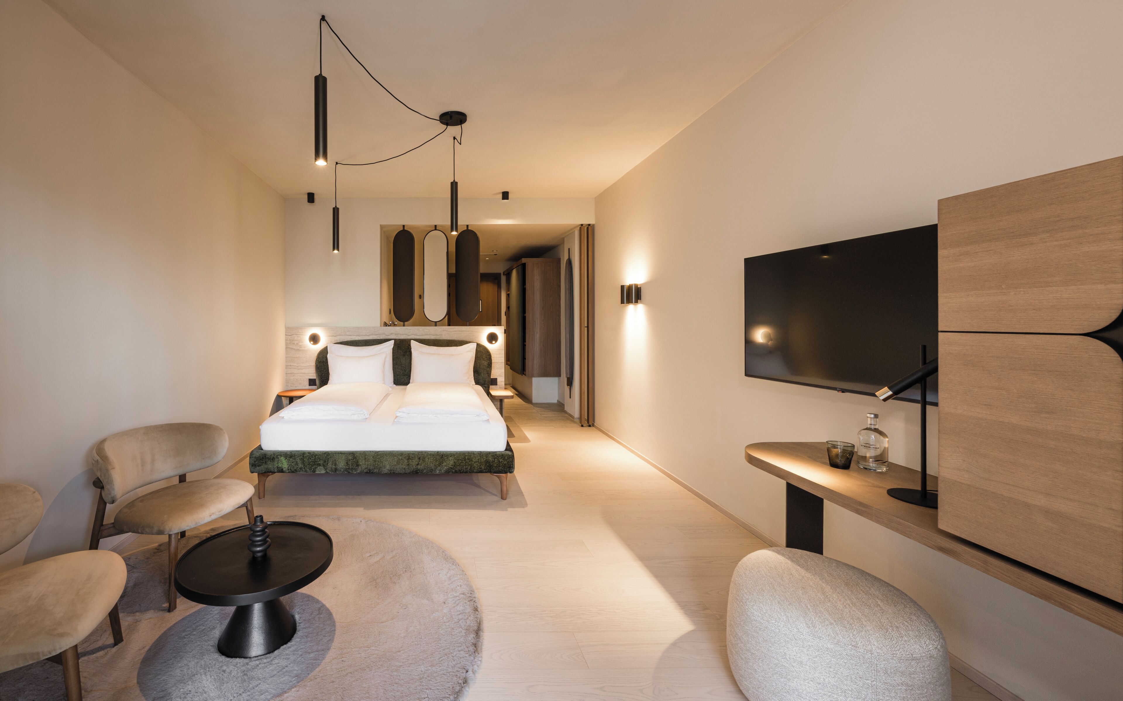 Quarto de hotel moderno com cama, duas cadeiras, uma mesinha, televisão e espelho.