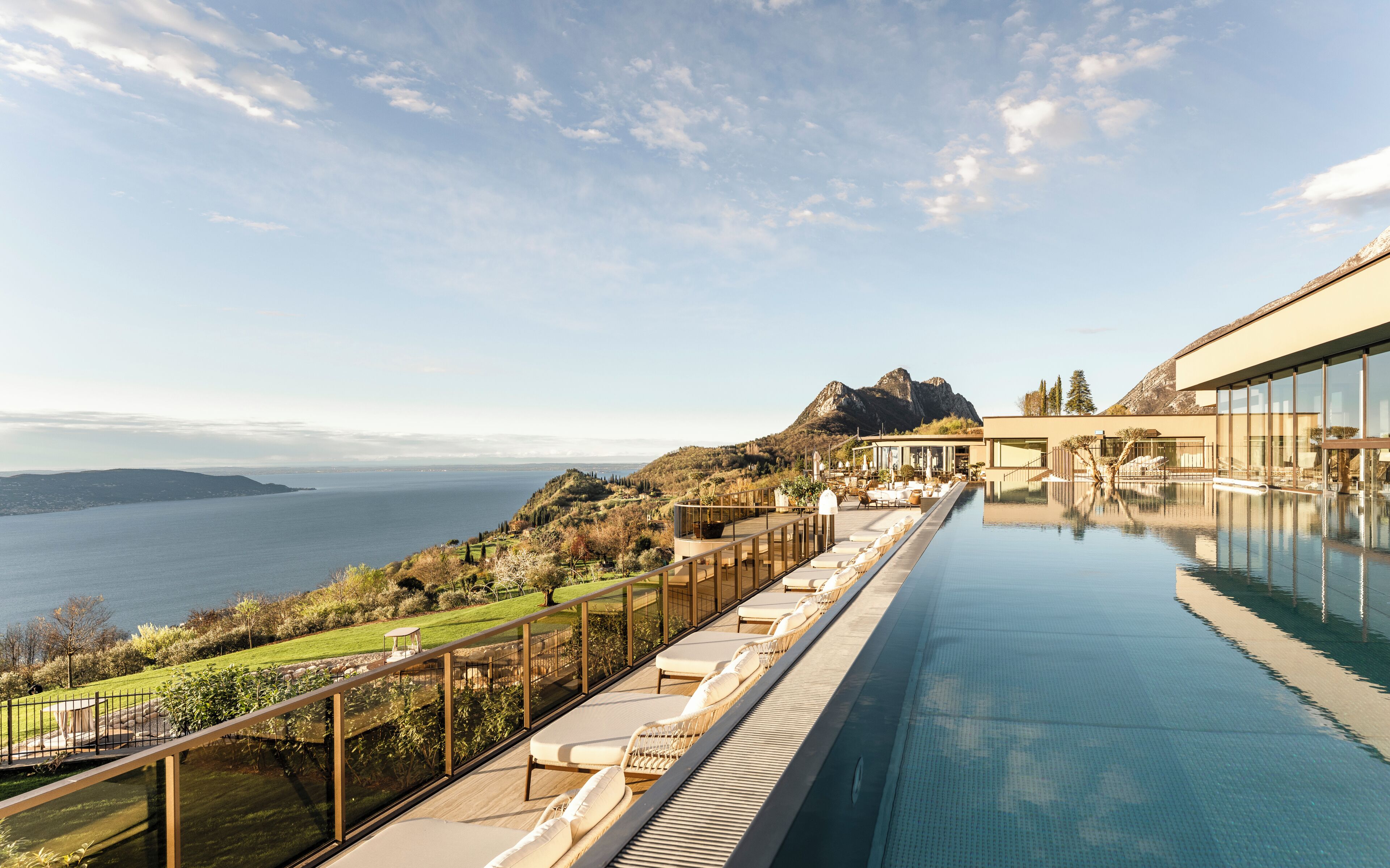 A pool with a view of a lake and mountains, a deck with lounge chairs, and a modern building with large windows.