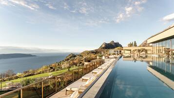 Ein Pool mit Blick auf einen See und Berge, eine Terrasse mit Liegestühlen und ein modernes Gebäude mit großen Fenstern.