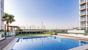 En moderne swimmingpool med fliseterrasse, omgivet af bygninger og en klar himmel.