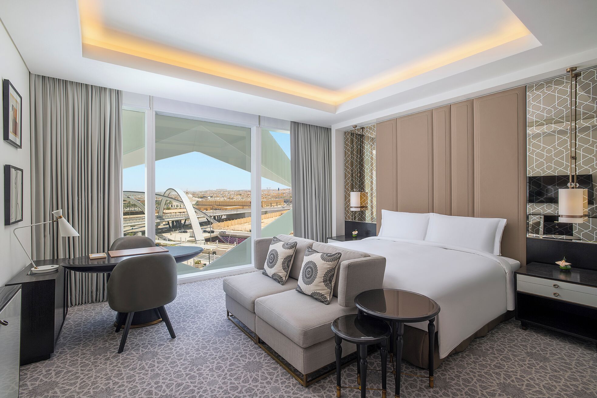 A modern hotel room with a large bed, a desk, a sofa, and a view of a cityscape through large windows.