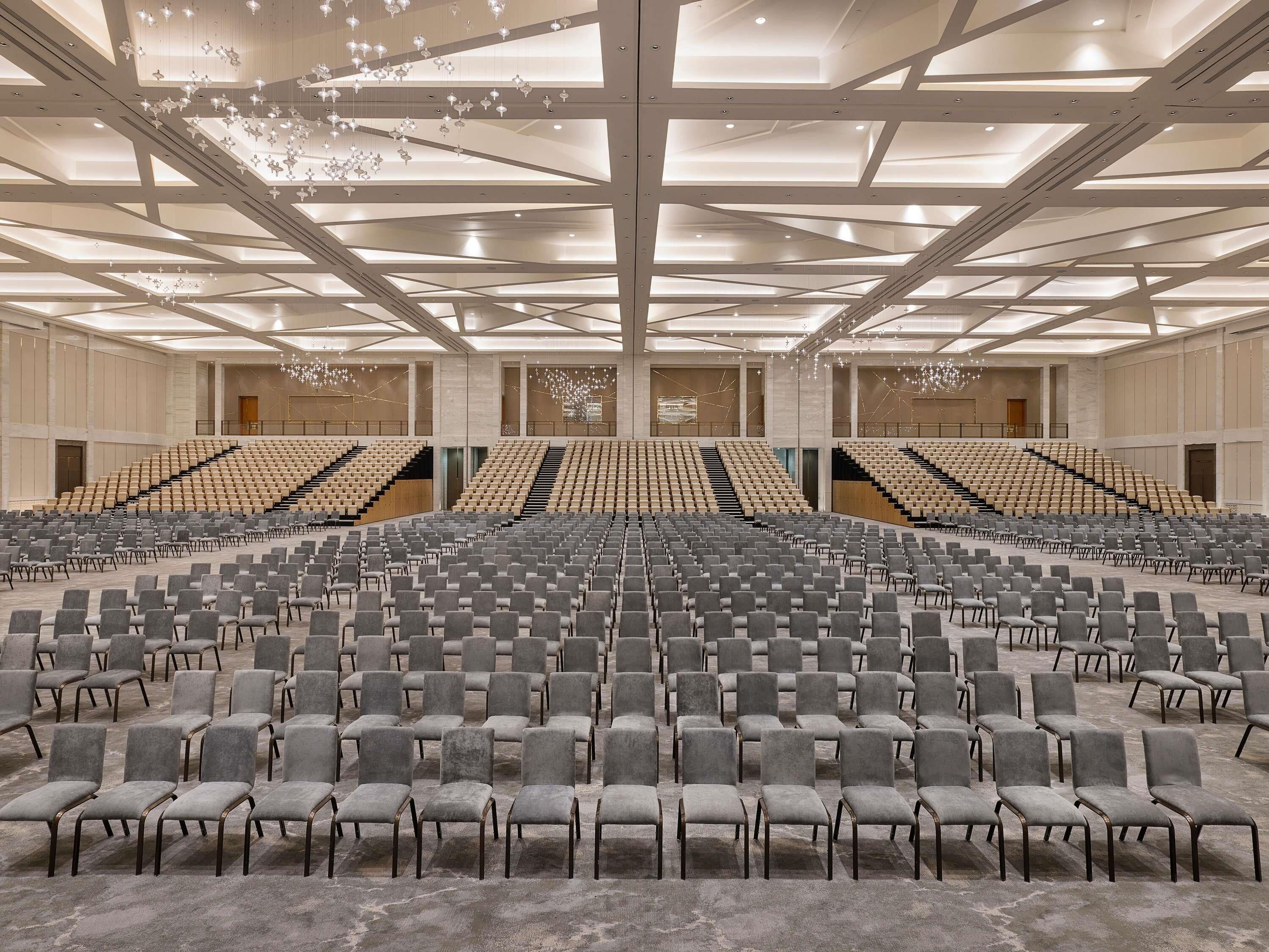 Ein großes, modernes Auditorium mit terrassenförmig angeordneten Sitzplätzen, einer hohen Decke mit geometrischen Mustern und einer neutralen Farbgestaltung.