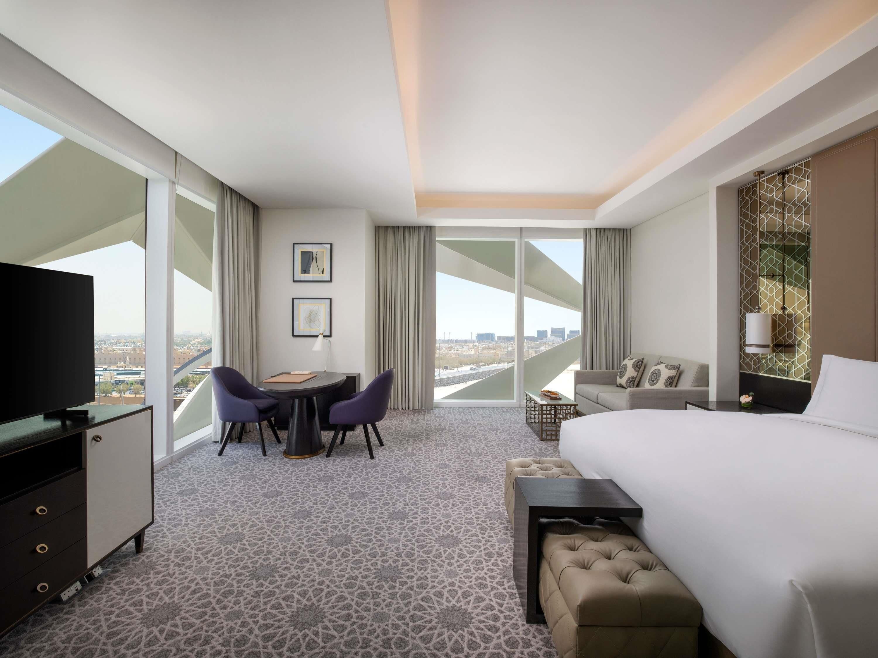 A hotel room with a bed, a desk with two chairs, a television, and a large window with a city view.