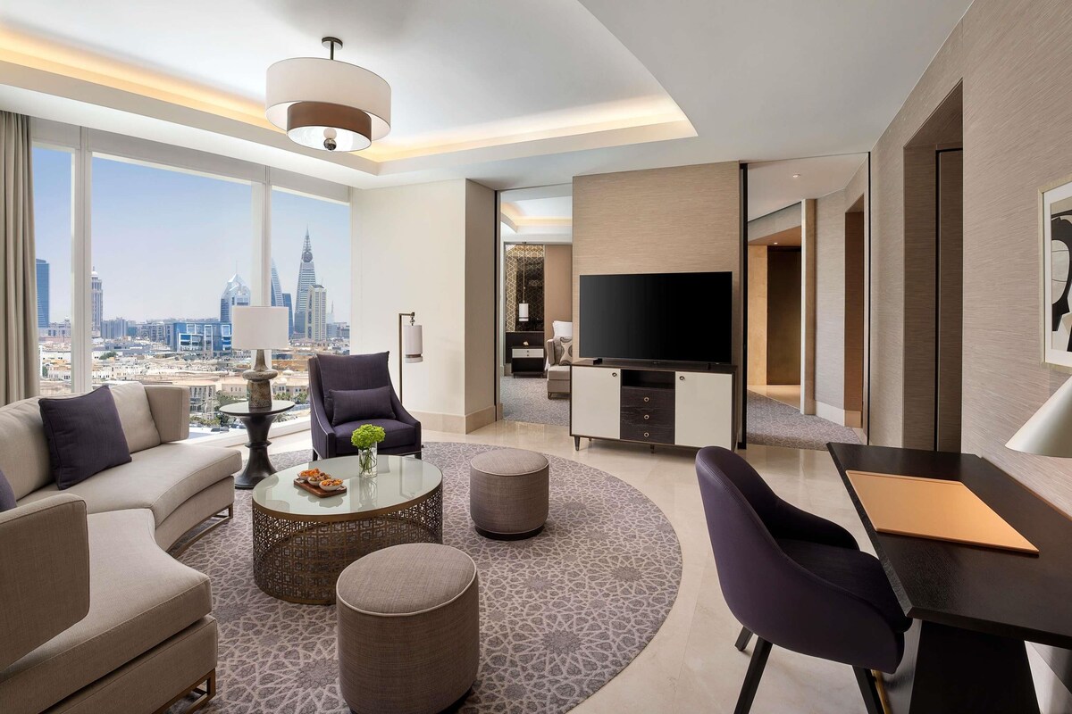 Una habitación de hotel moderna con sofá, sillón, otomanas y una mesa de centro redonda, además de un televisor de pantalla plana.
