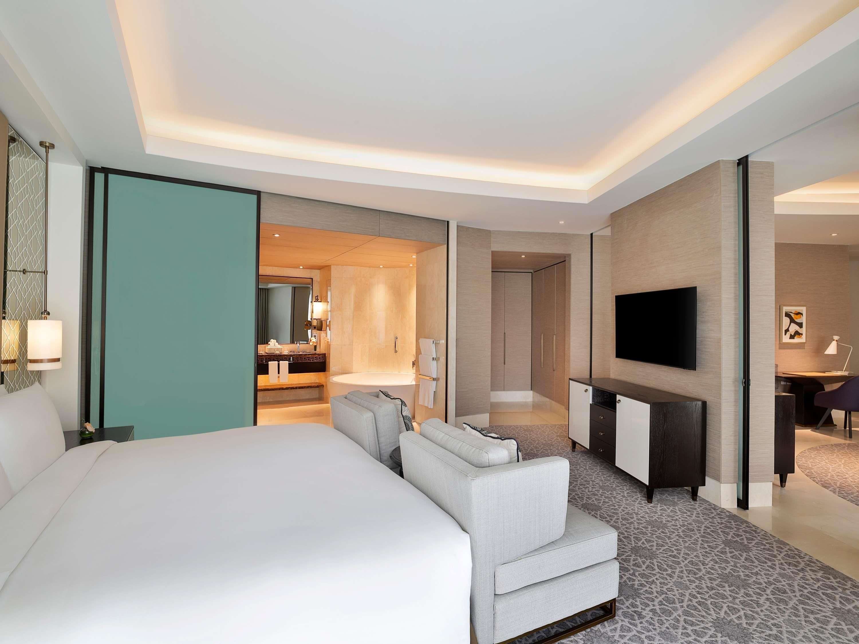 A modern hotel room with a large bed, a seating area, a TV, and a bathroom visible through an open door.
