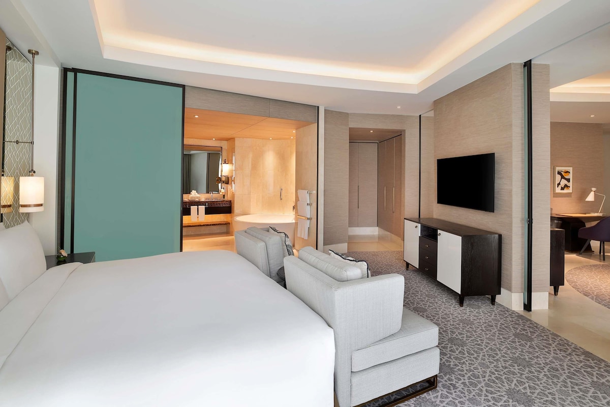 A modern hotel room with a large bed, a seating area, a TV, and a bathroom visible through an open door.