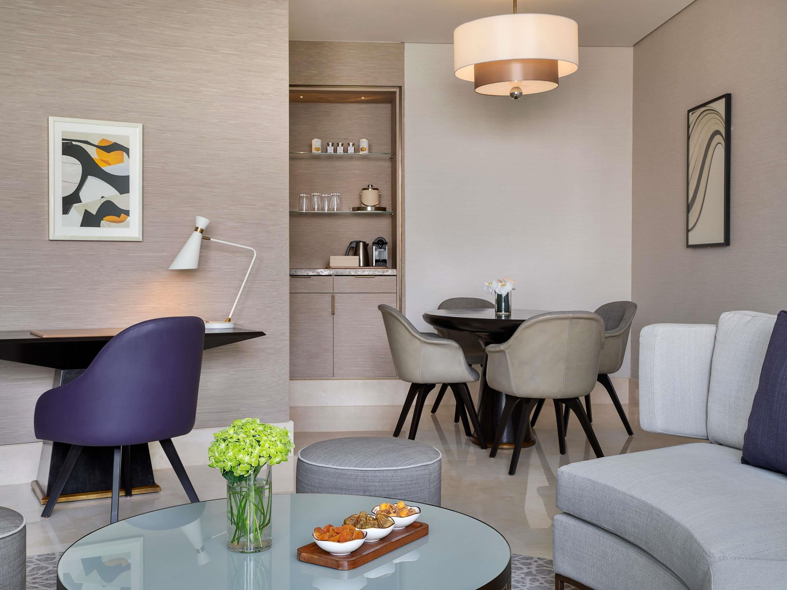 A modern living room with a glass coffee table, a purple chair, a grey sofa, a dining area with a round table and chairs, and a kitchen area with cabinets and a countertop.