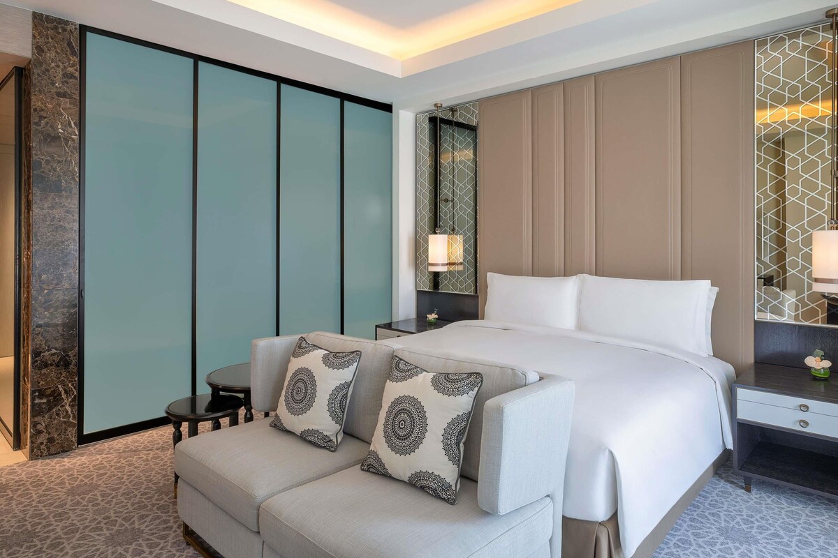 Habitación de hotel con cama, sofá, mesita de noche, lámpara y un panel decorativo en la pared.