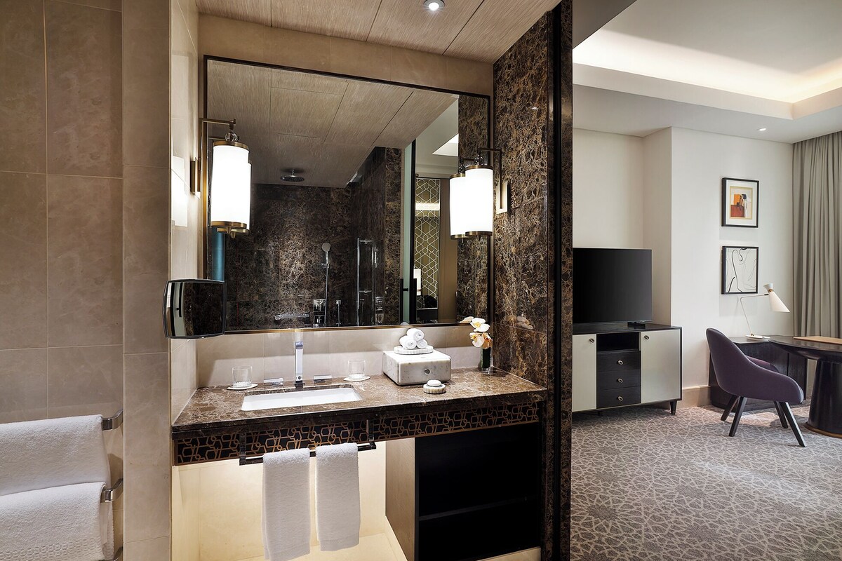 A modern bathroom with a large mirror, a sink, and a shower. There is a vanity with a mirror and a television in the corner.