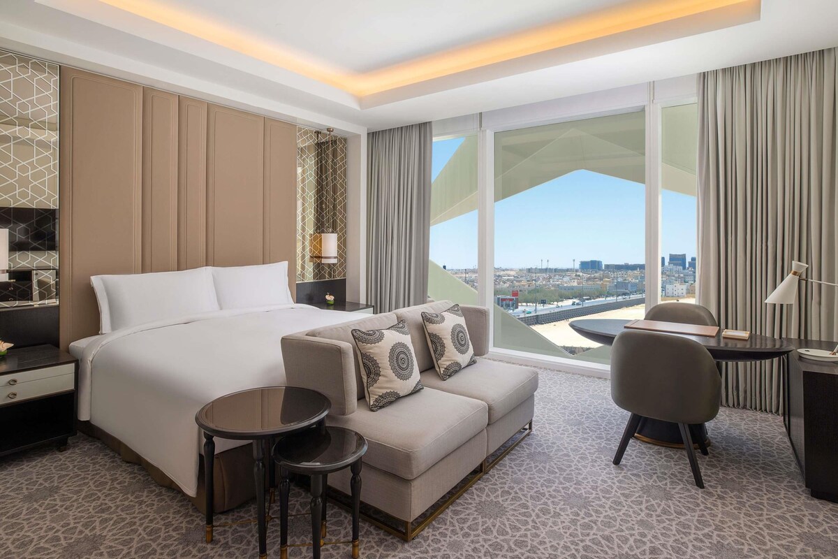 Una habitación de hotel moderna con una cama grande, un sofá, dos mesitas, un escritorio y vistas a la ciudad.