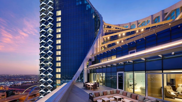 A modern hotel with a curved glass facade, outdoor seating area, and a view of the cityscape.