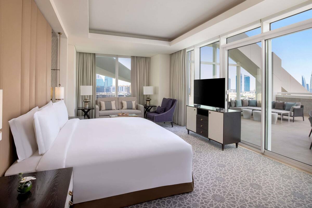 A hotel room with a large bed, a desk with a TV, a sofa, and a balcony with a city view.