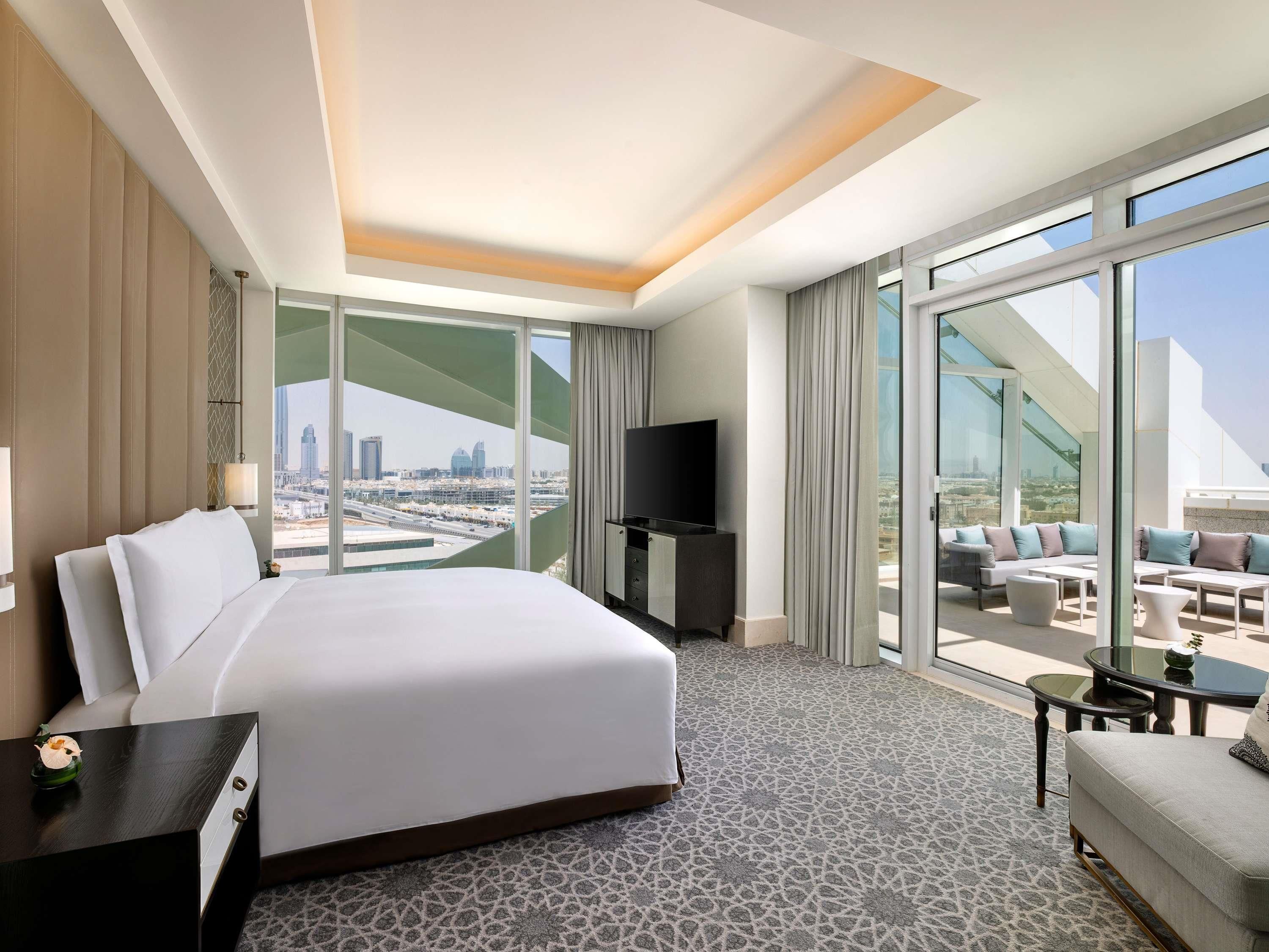 A hotel room with a large bed, a TV, a balcony with seating, and a city view.