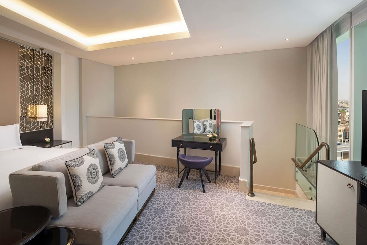 Ein Hotelzimmer mit Bett, einer Sitzecke, einem Schreibtisch, einem Stuhl, einem Spiegel und Blick auf die Stadt durch ein Fenster.