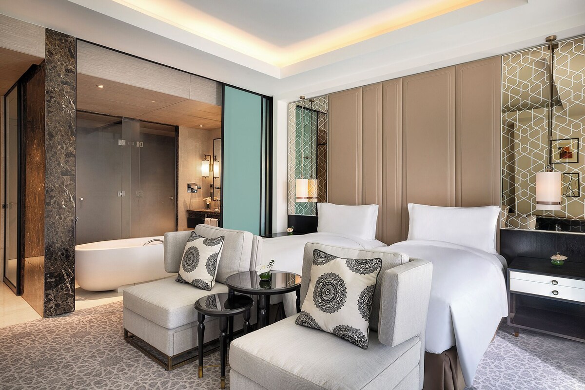 A hotel room with a large bed, two armchairs, a small table, a bathtub, and a vanity with a mirror.