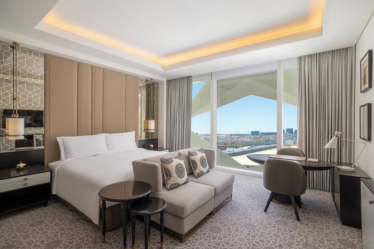 A modern hotel room with a large bed, a sofa, a dining table, and a view of the city.