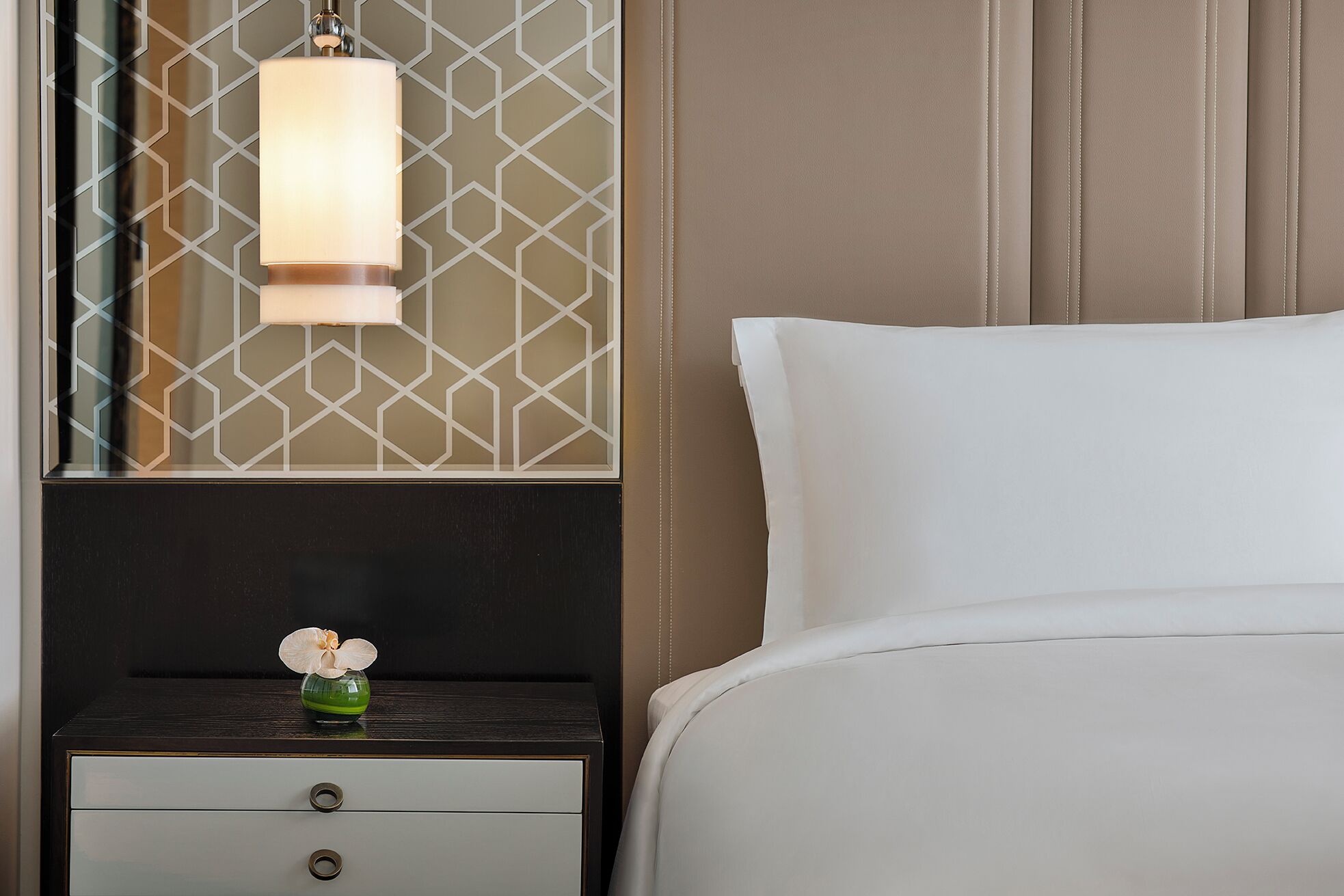 A neatly made bed with white linens, a bedside table with a small vase of flowers, and a wall-mounted lamp.