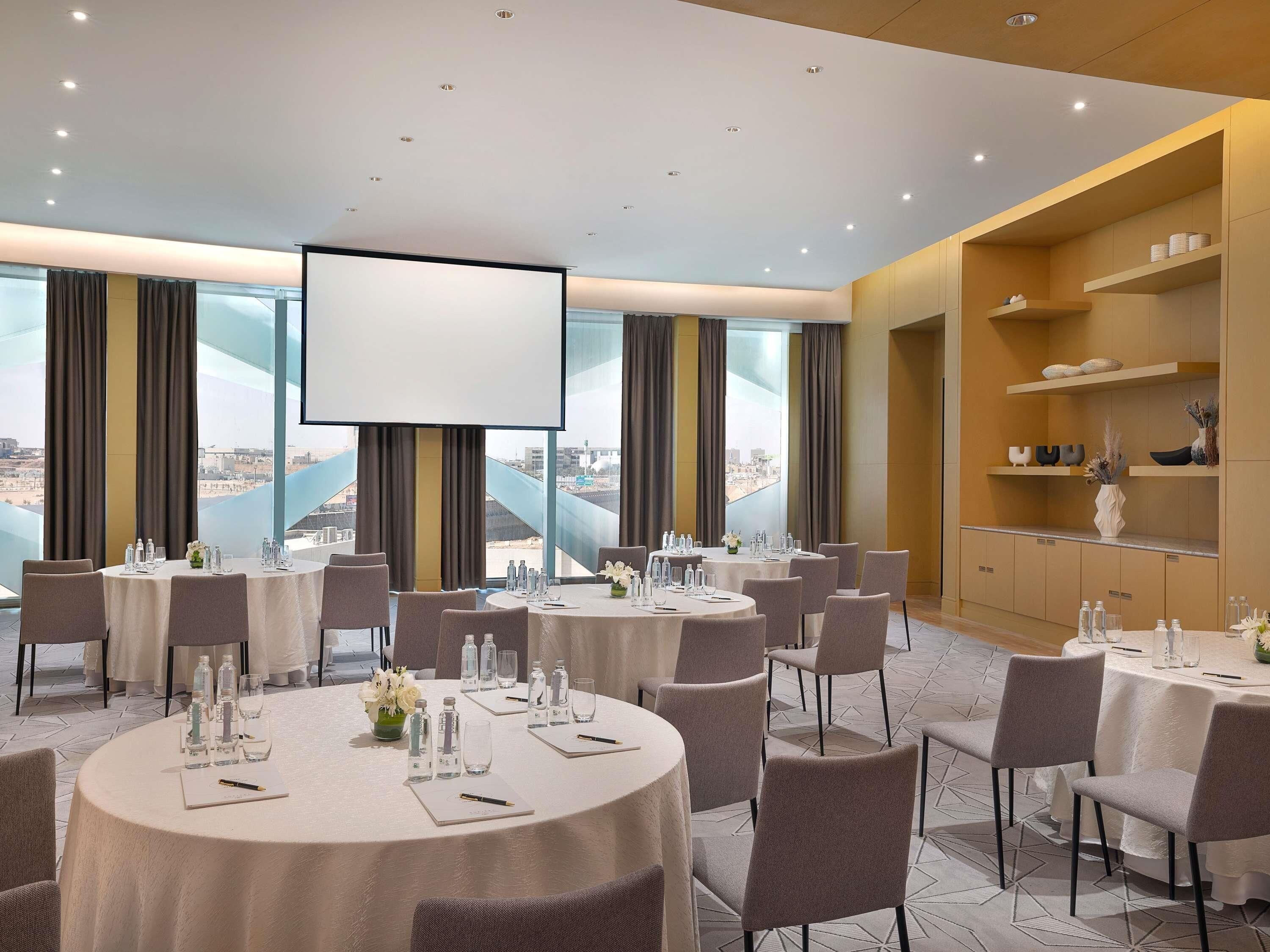 A conference room with multiple tables, chairs, a large screen, and a view of a cityscape through large windows.