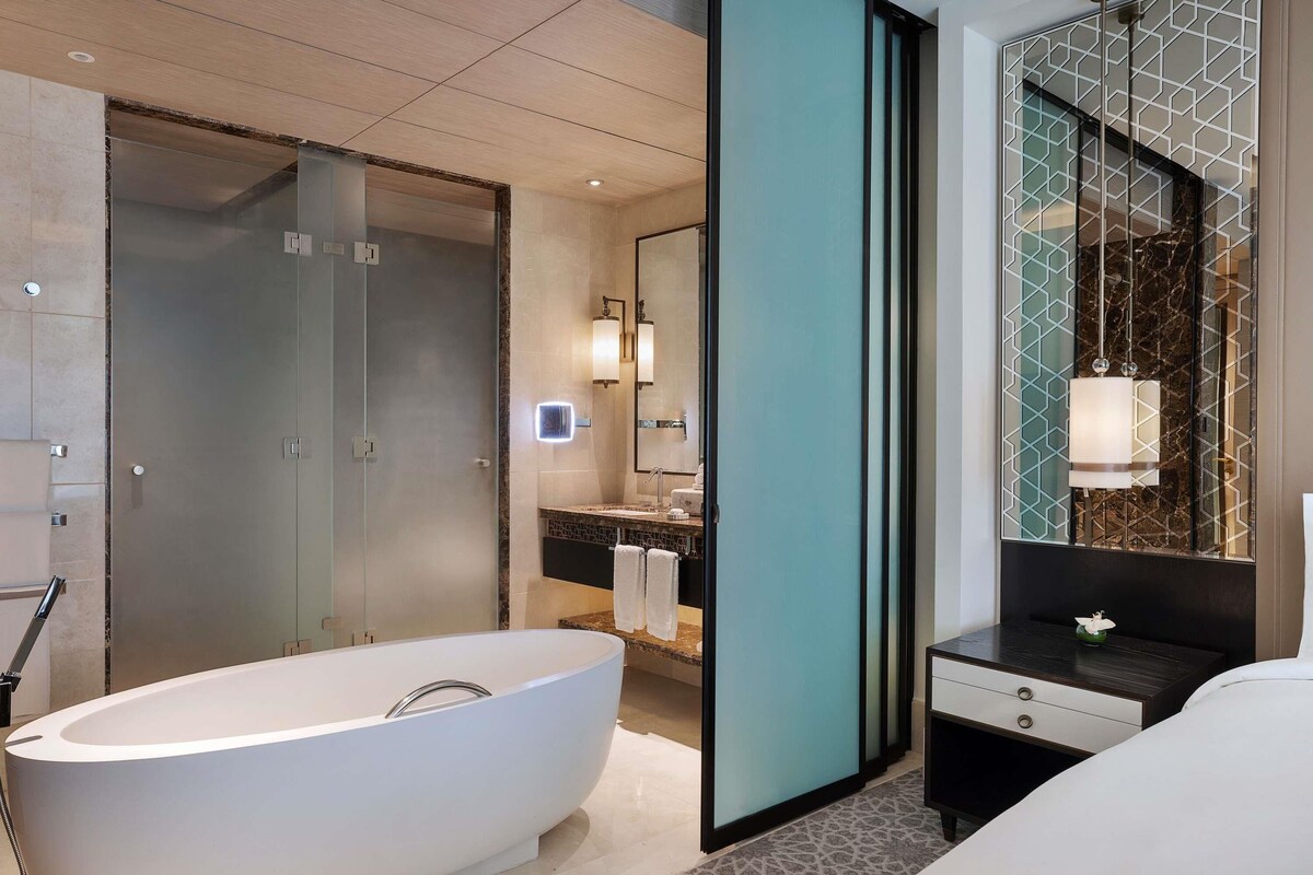 A modern bathroom with a freestanding bathtub, a vanity with a mirror, and a nightstand with a lamp.