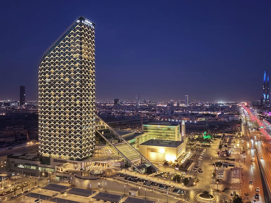 Um hotel moderno com fachada geométrica marcante, um pátio central e vista para o horizonte urbano noturno.