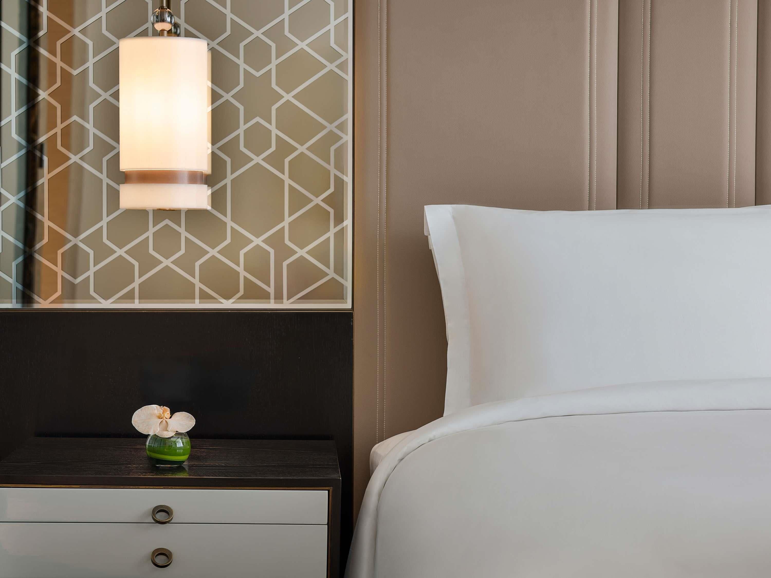 A neatly made bed with white linens, a bedside table with a dark wood finish, a vase with a single orchid, and a geometric patterned wall.
