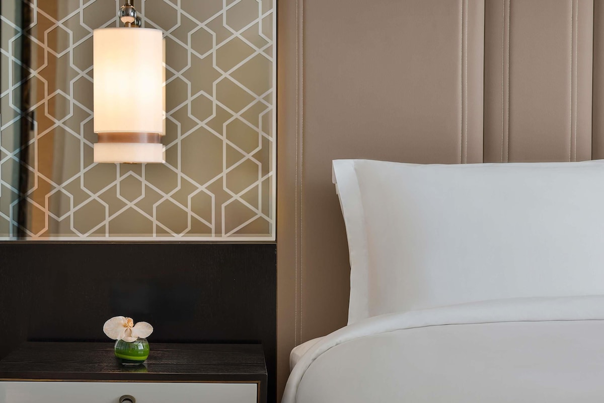 A neatly made bed with white linens, a bedside table with a dark wood finish, a vase with a single orchid, and a geometric patterned wall.