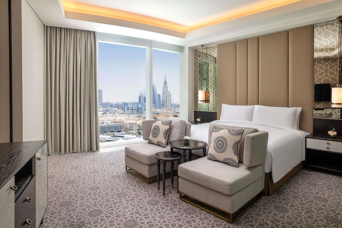 Ein Hotelzimmer mit einem Bett, zwei Sesseln, einem kleinen Tisch, einem Nachttisch und einem großen Fenster mit Blick auf die Stadt.