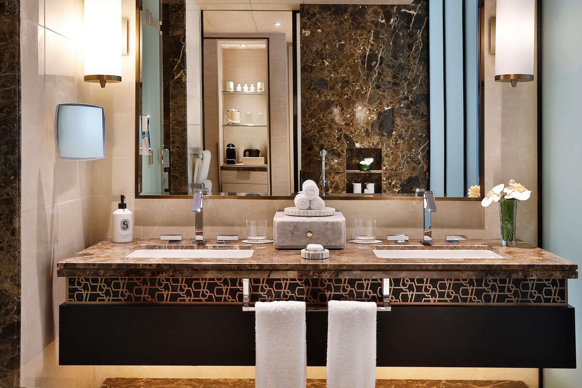 Une salle de bain moderne avec un meuble-lavabo en marbre, deux vasques, un grand miroir et un vase rempli de fleurs.