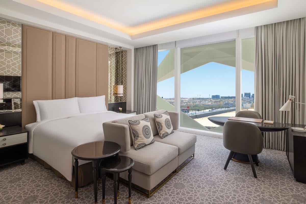 Ein modernes Hotelzimmer mit einem großen Bett, einem Sofa, zwei kleinen Tischen, einem Schreibtisch und einem großen Fenster mit Blick auf die Stadt.