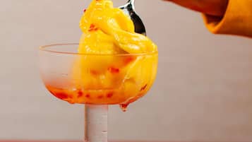 Un tazón de vidrio transparente con un postre amarillo con motas rojas, sostenido por una cuchara.