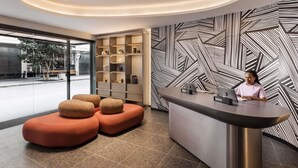 Un espace d’accueil au design moderne, comprenant un comptoir d’enregistrement, un coin salon avec des poufs orange, un mur à motifs et une porte vitrée donnant sur l’extérieur.