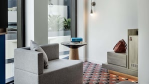 Une chambre d’hôtel moderne avec un canapé gris, une petite table ronde sur laquelle repose un livre, un lampadaire fixé au mur et un tapis à motifs.