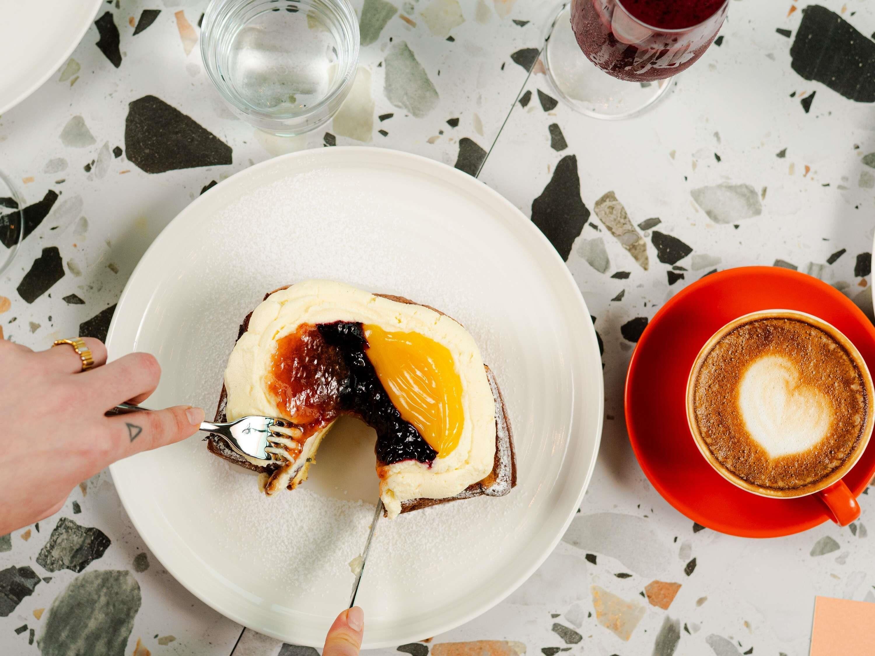 Una fetta di pane tostato con marmellata e un cucchiaio di burro su un piatto, una tazza rossa con un disegno a cuore nel latte e un bicchiere di bevanda rossa su un tavolo screpolato.