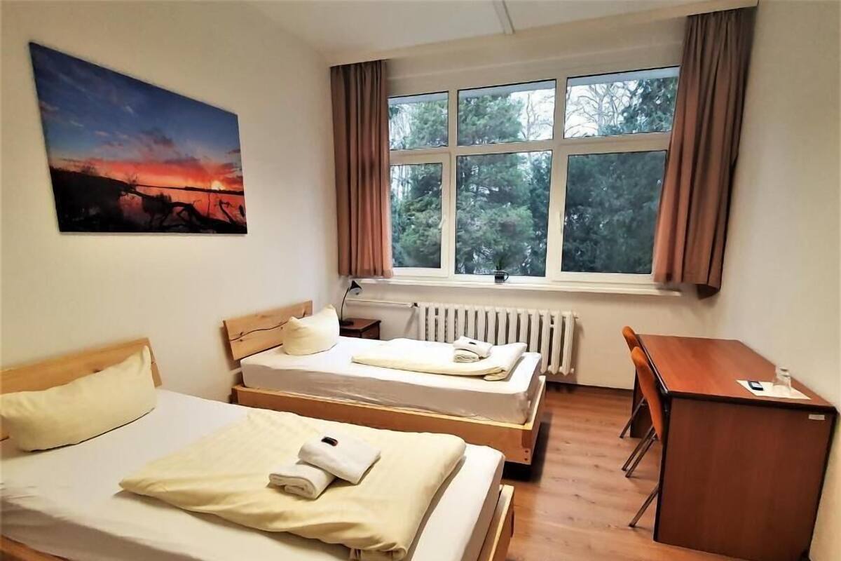 Une chambre avec deux lits simples, un bureau en bois, un radiateur et une fenêtre avec des rideaux.