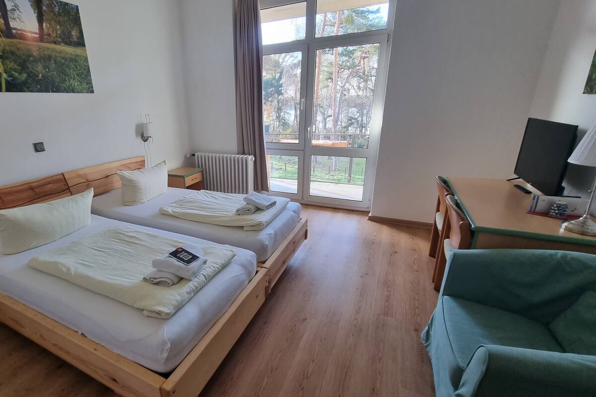 Une chambre d’hôtel avec deux lits, un bureau, une chaise, une télévision et une fenêtre donnant sur des arbres.