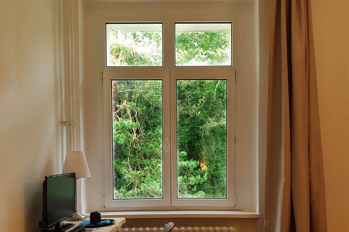 Une chambre à coucher avec un lit, un bureau avec un ordinateur, une fenêtre avec des rideaux et une vue sur la verdure à l’extérieur.