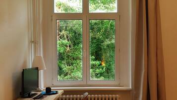 Ein Schlafzimmer mit einem Bett, einem Schreibtisch mit Computer, einem Fenster mit Vorhängen und Blick auf Grünflächen.