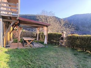 Eine Hollaube mit Tisch und Stühlen, ein Steinofen und Blick auf einen bewaldeten Hügel.