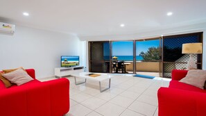 A modern living room with red sofas, a white coffee table, and a TV displaying a beach scene. - Stunning Oceanfront Retreat – Your Perfect Holiday Escape! (Gold Coast)