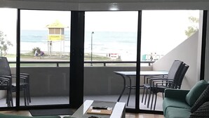 Une pièce avec un canapé vert, des chaises en bois, une table et un ventilateur de plafond. De grandes fenêtres offrent une vue sur la plage et sur le poste de surveillance des sauveteurs.