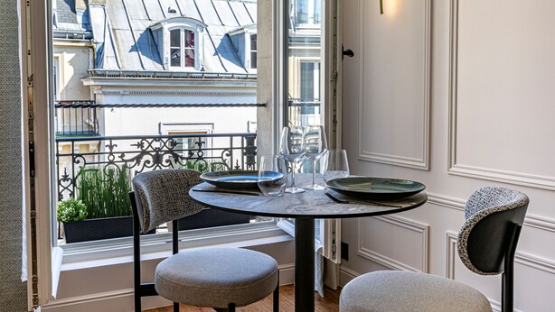 Comedor con una mesa redonda, dos sillas y vistas a un balcón con plantas y edificios.