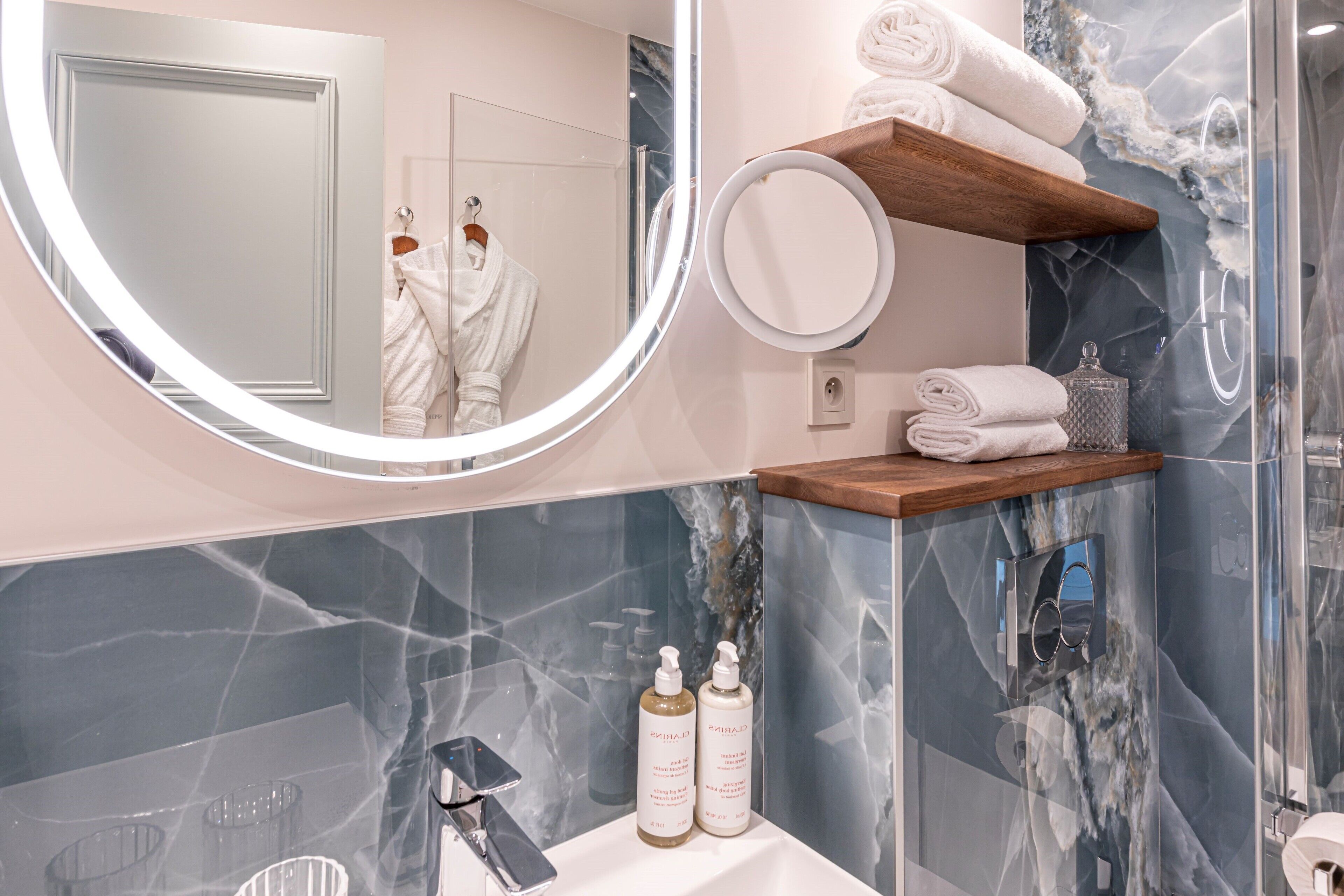 Un baño moderno con un revestimiento de mármol, un espejo redondo, un estante de madera con toallas y dos botellas de artículos de aseo.
