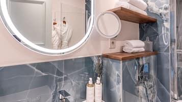 Un baño moderno con un revestimiento de mármol, un espejo redondo, un estante de madera con toallas y dos botellas de artículos de aseo.