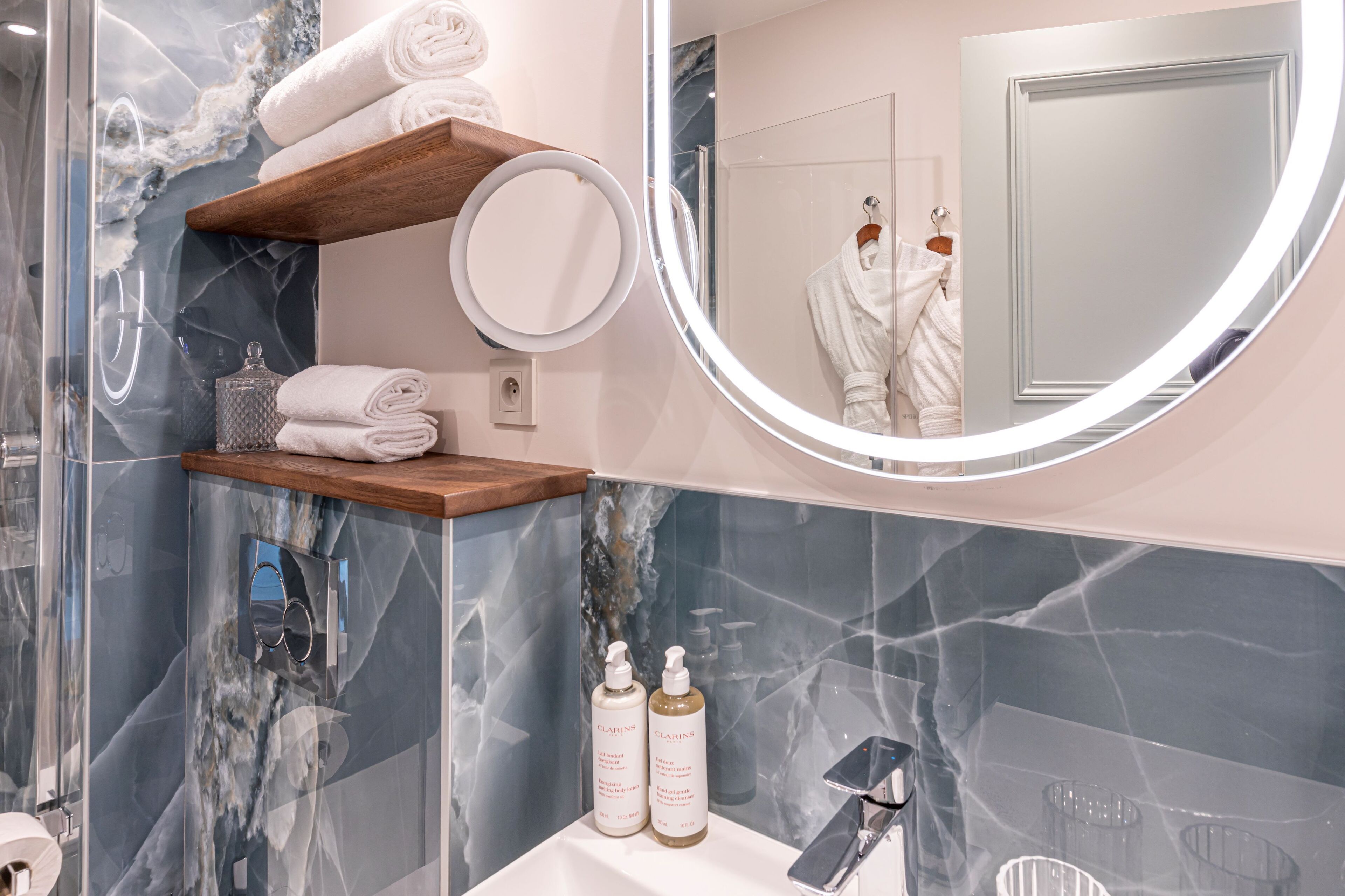 A bathroom with a marble backsplash, a wooden shelf with towels, a round mirror, and two bottles of lotion.