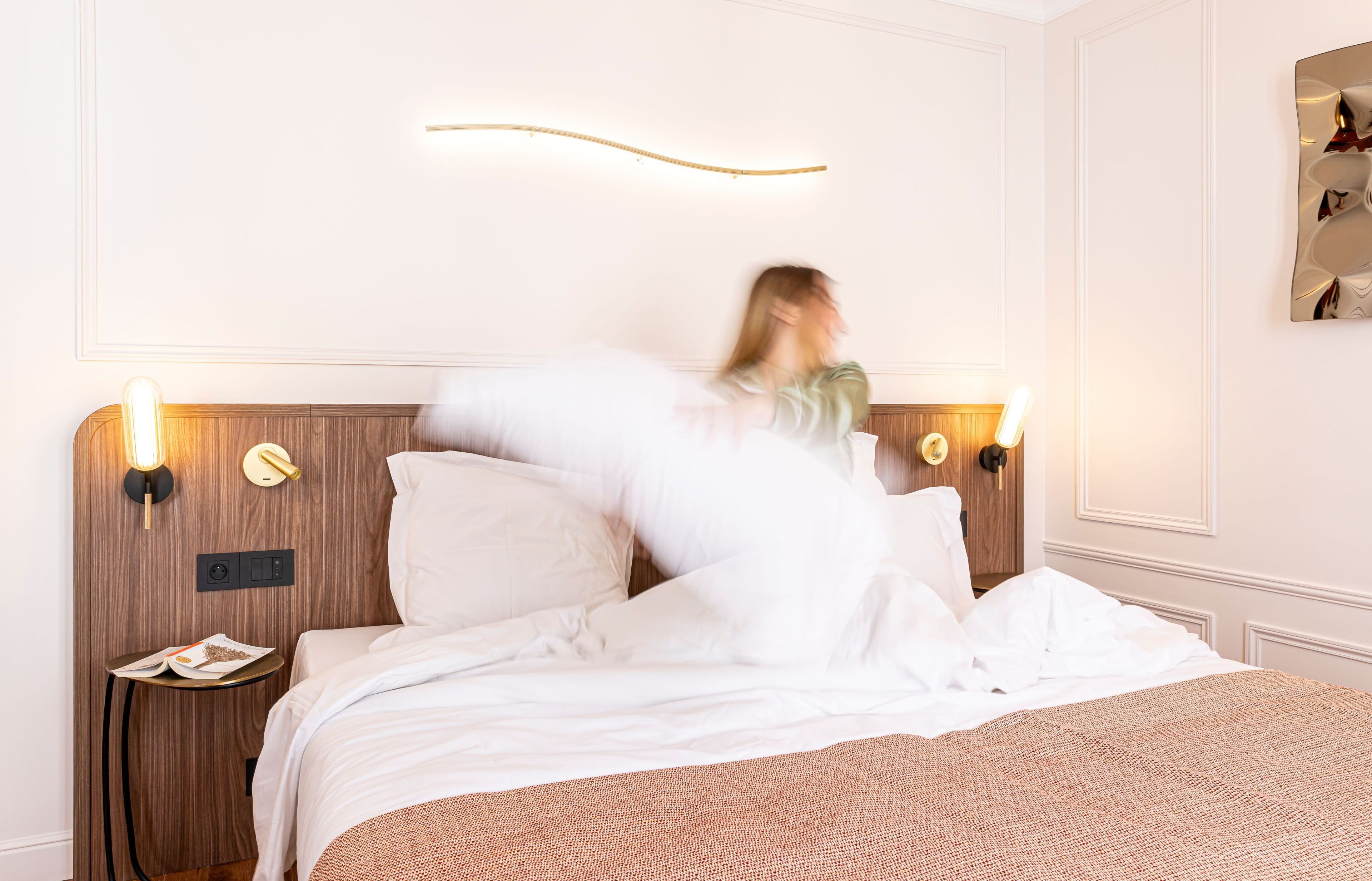 A hotel room with a wooden headboard, bedside lamps, a small table with a book, and a wall-mounted light fixture.