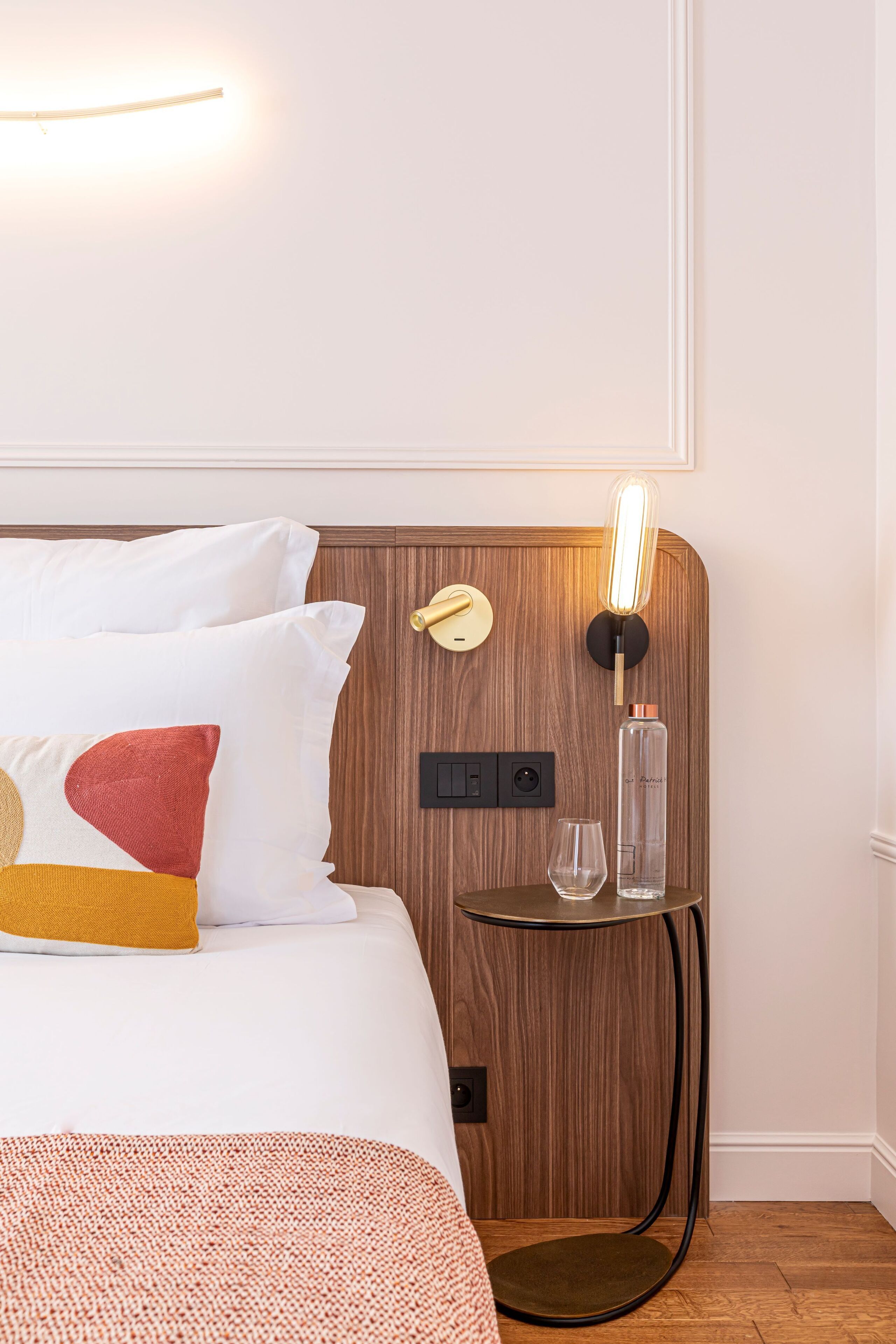 A neatly made bed with white and multicolored pillows, a small round table with a glass and a bottle, and a wall-mounted light fixture.