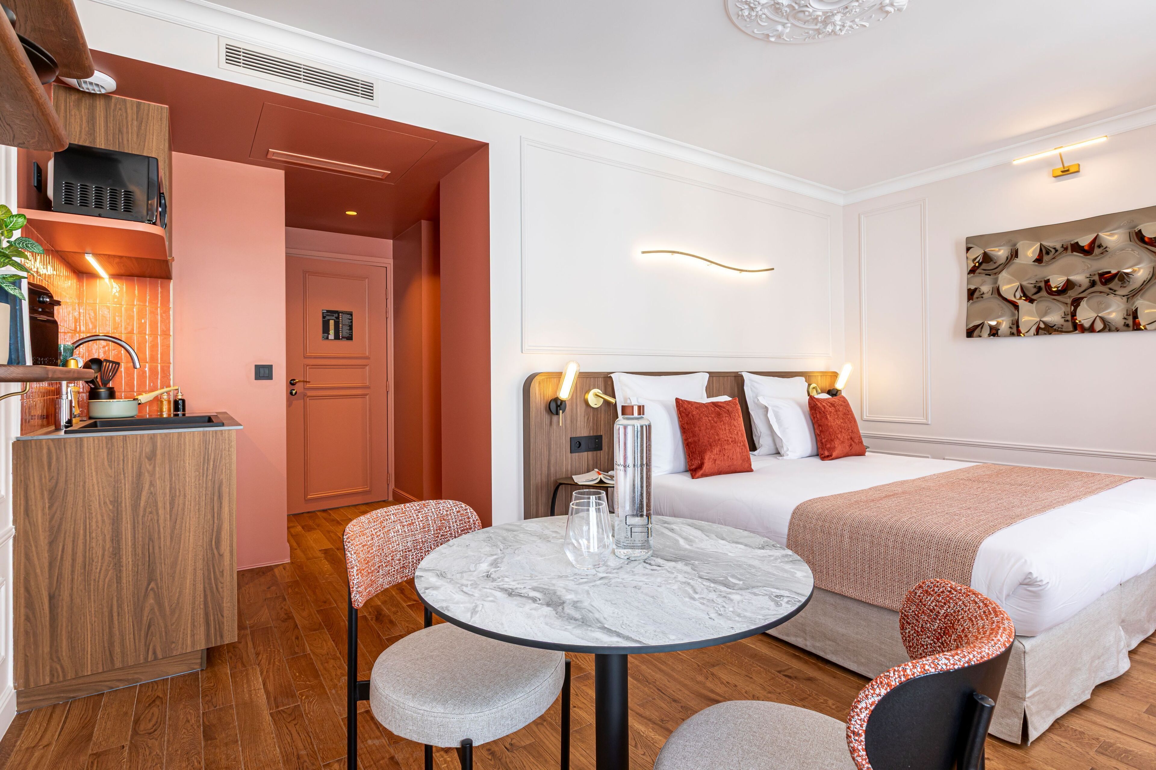 Una habitación de hotel moderna con una kitchenette de madera, una mesa redonda de mármol con dos sillas, una cama con colcha blanca y naranja, y una obra de arte abstracto colgada en la pared.